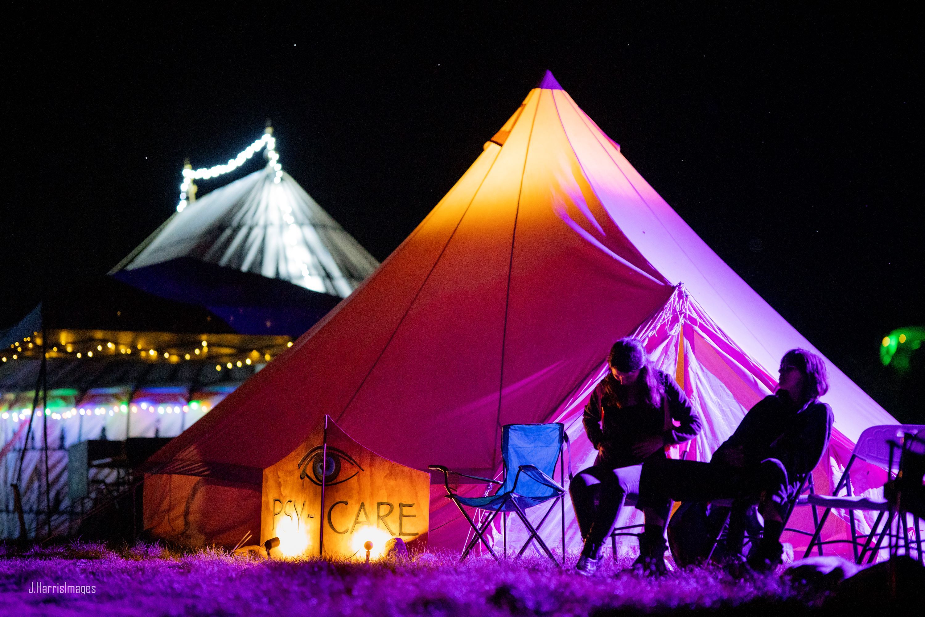 a night-time shot of the Psy-Care space, a bell-tent is in the background and lit warmly from inside. In front of it are a couple of people in pink hi-vis resting in camping chairs, with the 'Psy-Care' sign lit up next to them.
